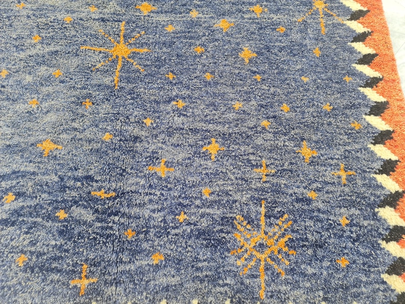 May include: A blue rug with a pattern of yellow stars and starbursts. The rug has a textured surface and a border with orange, black, and white geometric designs. The rug is likely used for home decor.