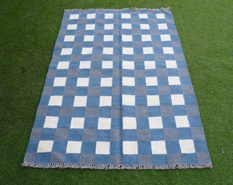 May include: A rectangular rug with a chequered pattern of blue, grey, and white squares. The rug is made of woven fabric and has a fringed edge. The rug is displayed on a green surface.