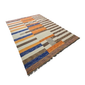 May include: A rectangular area rug with a geometric pattern. The rug features stripes and blocks in shades of orange, brown, blue, and gray against a cream background. The rug has a fringed edge.