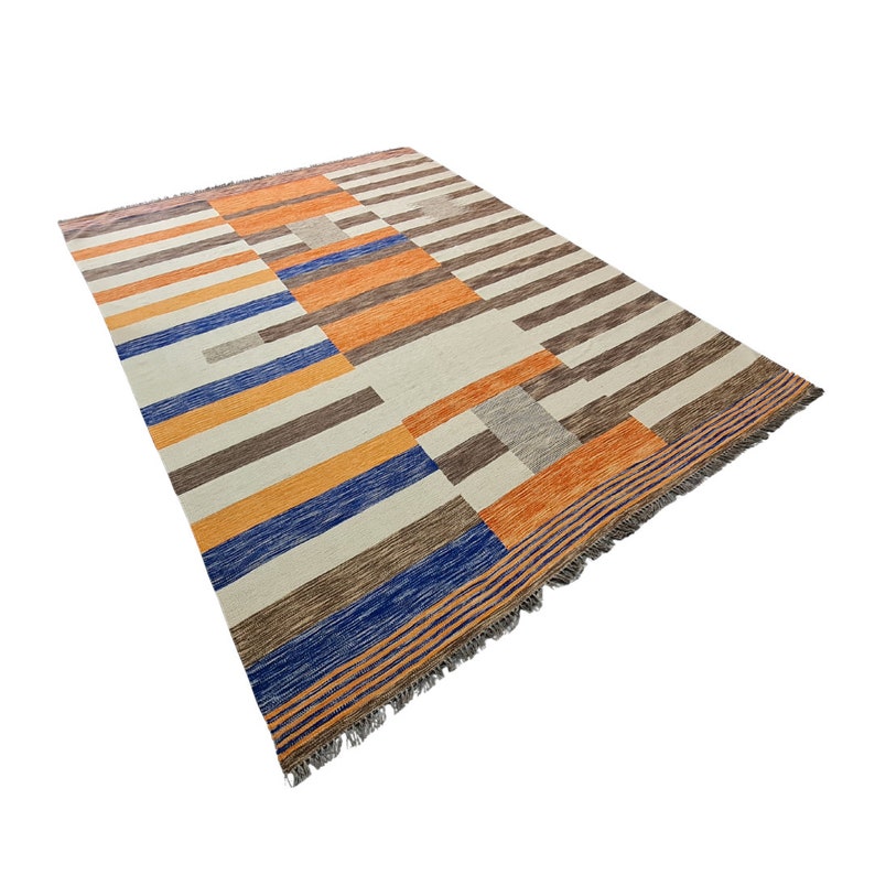 May include: A rectangular area rug with a geometric pattern. The rug features stripes and blocks in shades of orange, brown, blue, and gray against a cream background. The rug has a fringed edge.