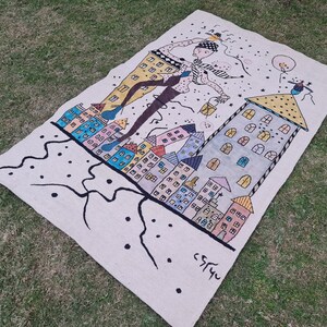 Op de afbeelding: Een rechthoekig, gebroken wit tapijt met een grillige illustratie van een figuur die boven kleurrijke gebouwen zweeft. Het kunstwerk heeft een zwarte omtrek en accenten van geel, blauw en roze. Het tapijt ligt op groen gras.
