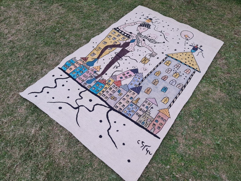 Op de afbeelding: Een rechthoekig, gebroken wit tapijt met een grillige illustratie van een figuur die boven kleurrijke gebouwen zweeft. Het kunstwerk heeft een zwarte omtrek en accenten van geel, blauw en roze. Het tapijt ligt op groen gras.
