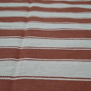 May include: A woven rug with alternating horizontal stripes of light beige and reddish-brown. The rug's texture appears slightly uneven, with visible variations in the weave. The stripes are of varying widths, creating a simple, yet visually appealing pattern.