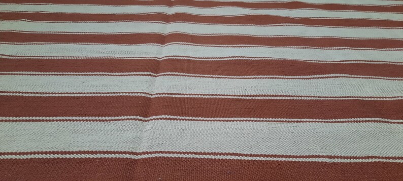 May include: A woven rug with alternating horizontal stripes of light beige and reddish-brown. The rug's texture appears slightly uneven, with visible variations in the weave. The stripes are of varying widths, creating a simple, yet visually appealing pattern.