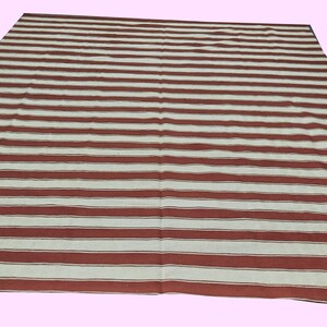 May include: A large rectangular rug with a pattern of alternating wide stripes in red and off-white. The rug is laid out on a light pink surface, and the stripes run horizontally across the rug.