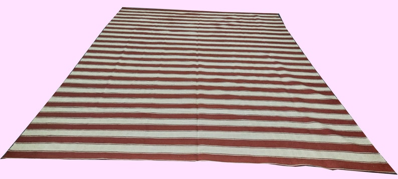 May include: A large rectangular rug with a pattern of alternating wide stripes in red and off-white. The rug is laid out on a light pink surface, and the stripes run horizontally across the rug.