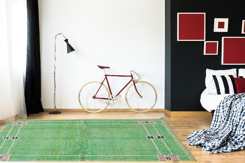 May include: A room with a green rug, a burgundy bicycle, and a black and white striped bedspread. A black floor lamp with a red cord stands near the bicycle. Red framed artwork hangs on a black wall.