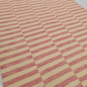 May include: A large rectangular rug with a repeating pattern of alternating red and beige horizontal stripes. The rug is made of a woven material and has a textured appearance. The stripes are evenly spaced and create a geometric design.