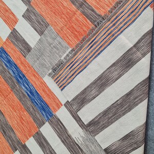 May include: Geometric patterned rug with a patchwork design. The rug features a combination of orange, blue, gray, and white stripes arranged in a diagonal pattern. The rug has a fringed edge and is made of woven fabric.