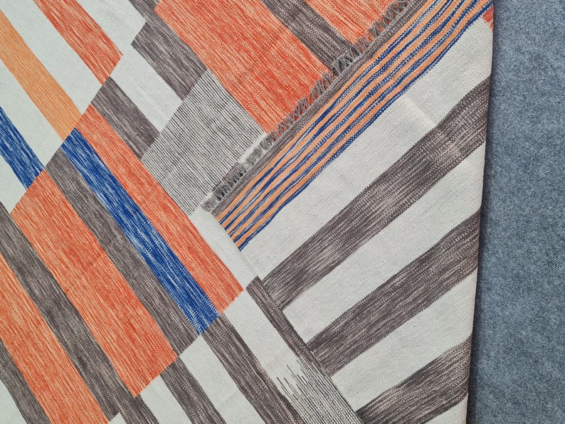 May include: Geometric patterned rug with a patchwork design. The rug features a combination of orange, blue, gray, and white stripes arranged in a diagonal pattern. The rug has a fringed edge and is made of woven fabric.