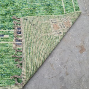 Puede incluir: Una alfombra tejida verde y blanca con un dise&ntilde;o geom&eacute;trico. La alfombra tiene una superficie texturizada y flecos en un borde. Se ve la parte inferior de la alfombra, mostrando un patr&oacute;n verde claro y beige. La alfombra est&aacute; sobre una superficie de hormig&oacute;n gris.