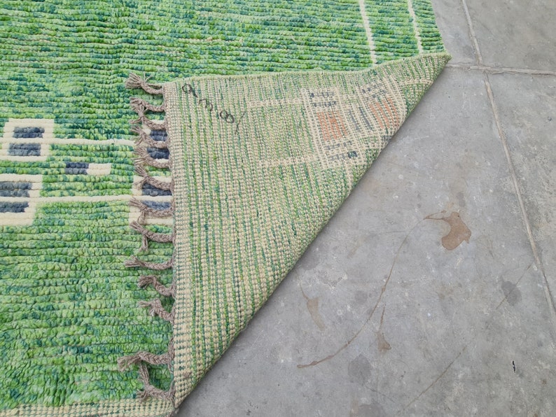 Puede incluir: Una alfombra tejida verde y blanca con un dise&ntilde;o geom&eacute;trico. La alfombra tiene una superficie texturizada y flecos en un borde. Se ve la parte inferior de la alfombra, mostrando un patr&oacute;n verde claro y beige. La alfombra est&aacute; sobre una superficie de hormig&oacute;n gris.