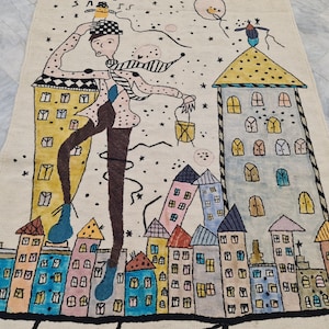 Op de afbeelding: Een grillig tapijt met een gestileerde stadsgezicht met kleurrijke gebouwen en een figuur met een hoed. Het ontwerp bevat sterren en een ballon, wat een speelse en artistieke esthetiek cre&euml;ert. Het tapijt is gemaakt van beige materiaal.