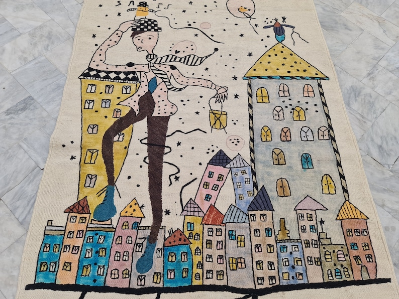 Op de afbeelding: Een grillig tapijt met een gestileerde stadsgezicht met kleurrijke gebouwen en een figuur met een hoed. Het ontwerp bevat sterren en een ballon, wat een speelse en artistieke esthetiek cre&euml;ert. Het tapijt is gemaakt van beige materiaal.