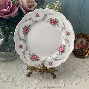 Puede incluir: Un plato de porcelana blanca con un delicado diseño floral con rosas rosas y volutas grises. El plato se muestra en un soporte de metal dorado.