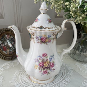 May include: White porcelain coffee pot with a floral design featuring pink, purple, and yellow roses. The pot has a gold trim and a decorative lid. The handle and spout are curved. The pot is on a white lace doily.