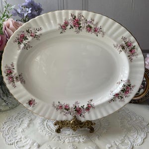 May include: A white oval serving platter with a gold rim and a floral pattern of pink roses and green leaves. The platter is on a white lace doily and a gold metal stand.