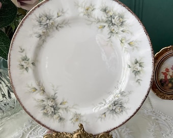 May include: A white porcelain plate with a gold rim and a floral design featuring white roses and green leaves. The plate is on a gold metal stand.