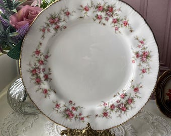 May include: A white porcelain dinner plate with a gold rim and a delicate floral pattern of pink roses. The plate is on a gold metal stand.