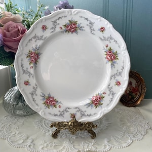 Op de afbeelding: Een wit porseleinen bord met een decoratief bloemmotief met roze en blauwe rozen. Het bord heeft een gegolfde rand en een grijs en wit bloemmotief rond de rand. Het bord wordt gepresenteerd op een gouden metalen standaard.