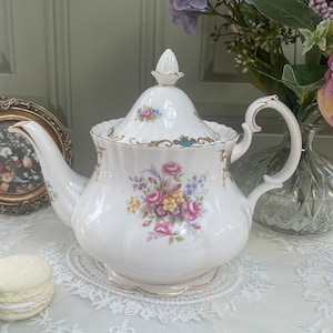 May include: White porcelain teapot with a floral design in pink, purple, and yellow. The teapot has a curved spout, a curved handle, and a lid with a decorative finial. Gold trim accents the edges. A classic tea party item.