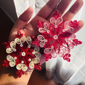 May include: Two handmade paper snowflakes, one red and white and one pink and red, are held in a hand. The snowflakes are made of rolled paper and have a delicate, intricate design.