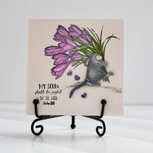 May include: A square ceramic tile featuring a watercolor illustration of a gray mouse holding purple flowers. The tile has the text "MY SOUL shall be joyful IN THE LORD Psalm 34". The tile is displayed on a black metal stand.