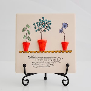 May include: A decorative square tile featuring three potted plants with green, blue, and purple flowers. The tile has the text "Nothing can separate us from the love of God which is in Christ our Lord Romans 8:38-39". The tile is displayed on a black metal stand.