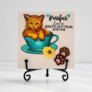 May include: A decorative square tile featuring a watercolor illustration of a ginger kitten sitting in a teal teacup. The tile includes the text "Purrfect love casts out fear" and a small sunflower and paw prints.