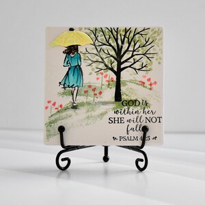 Puede incluir: Una baldosa de cerámica cuadrada con una ilustración de acuarela de una mujer con un paraguas amarillo caminando cerca de un árbol. La baldosa tiene el texto "God is within her, she will not fall. Psalm 4:5". La baldosa se muestra en un soporte de metal negro.