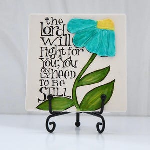 Könnte beinhalten: Eine quadratische Keramikfliese mit einem türkis-gelben Blumenmuster und den Worten "the lord will fight for you; you only need to be still" in schwarzer Schrift. Die Fliese wird auf einem schwarzen Metallständer präsentiert.