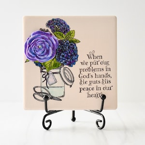Könnte beinhalten: Eine cremefarbene quadratische Fliese mit einer Aquarellillustration einer lila Rose und blauen Blumen in einem Glas. Die Fliese trägt den Text "When we put our problems in God's hands, He puts His peace in our heart."