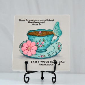 Könnte beinhalten: Dekorative Keramikfliese mit einem Design einer türkisfarbenen Tasse und Untertasse, verziert mit rosa Blumen und einem Schmetterling. Die Fliese enthält den Text "Do not let your hearts be troubled and do not be afraid John 14:27" und "I AM always with you Matthew 28:19-20".