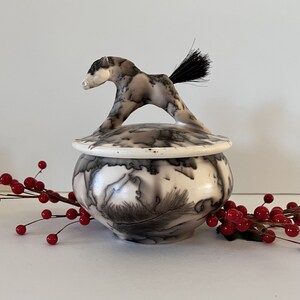 May include: A white ceramic jar with a black and white feather design. The lid is shaped like a horse with a black tail.