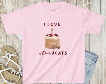 May include: A light pink t-shirt featuring the text "I LOVE JELLYCATS" in brown letters. The shirt has a graphic of a cake with a candle. The cake has a smiling face and is decorated with red berries.