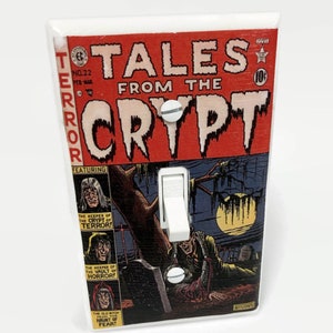 May include: A light switch cover featuring a vintage comic book cover design. The cover is titled "TALES FROM THE CRYPT" with a red background and illustrations of horror scenes. The switch is white and centered.