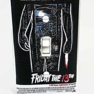 May include: A light switch cover featuring the "Friday the 13th" movie poster design. The black cover has a forest scene with two moons, a white switch, and the text "Friday the 13th".