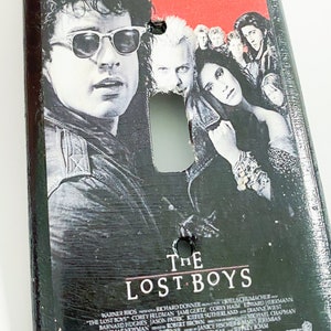 May include: A black light switch cover featuring the movie poster for "The Lost Boys." The poster art includes images of actors and the film's title. The cover has a distressed, textured finish.