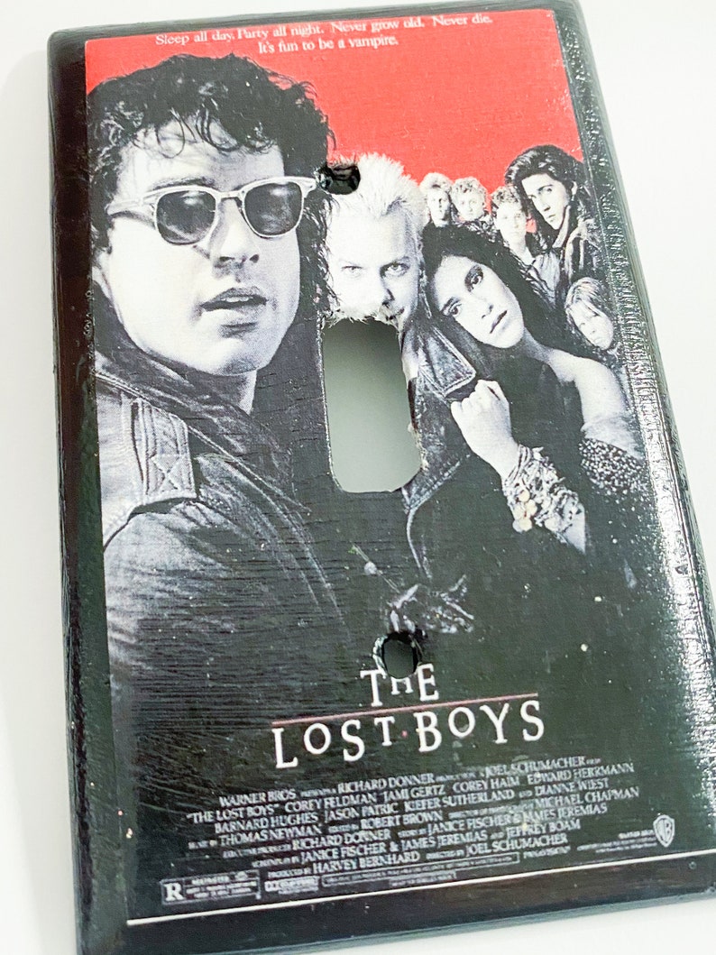 May include: A black light switch cover featuring the movie poster for "The Lost Boys." The poster art includes images of actors and the film's title. The cover has a distressed, textured finish.