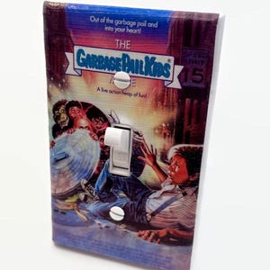 May include: A light switch cover featuring artwork from "The Garbage Pail Kids Movie." The cover has a colorful design with the movie title and characters. The switch is white, and the cover is secured with two screws.