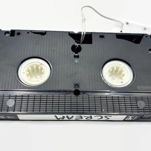 Może przedstawiać: Czarna kaseta VHS z napisem "SCREAM" na etykiecie. Taśma ma białe szpule i biały wieszak. Przedmiot w stylu retro.