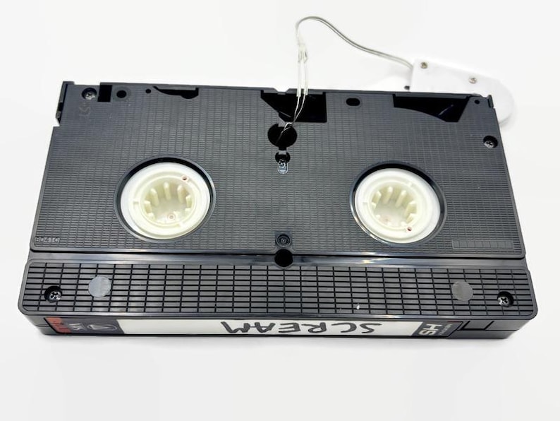 Może przedstawiać: Czarna kaseta VHS z napisem "SCREAM" na etykiecie. Taśma ma białe szpule i biały wieszak. Przedmiot w stylu retro.