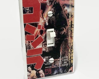 May include: A white light switch cover featuring a vintage Godzilla movie poster design. The poster has red Japanese text and a black and white image of Godzilla. The switch and screws are white.