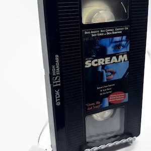 Może przedstawiać: Czarna kaseta VHS z filmem "Scream" z tekstem "Clever, Hip And Scary!" i nazwiskami aktor&oacute;w. Taśma jest trzymana przez srebrny druciany stojak i ma dołączony biały prostokątny przedmiot.