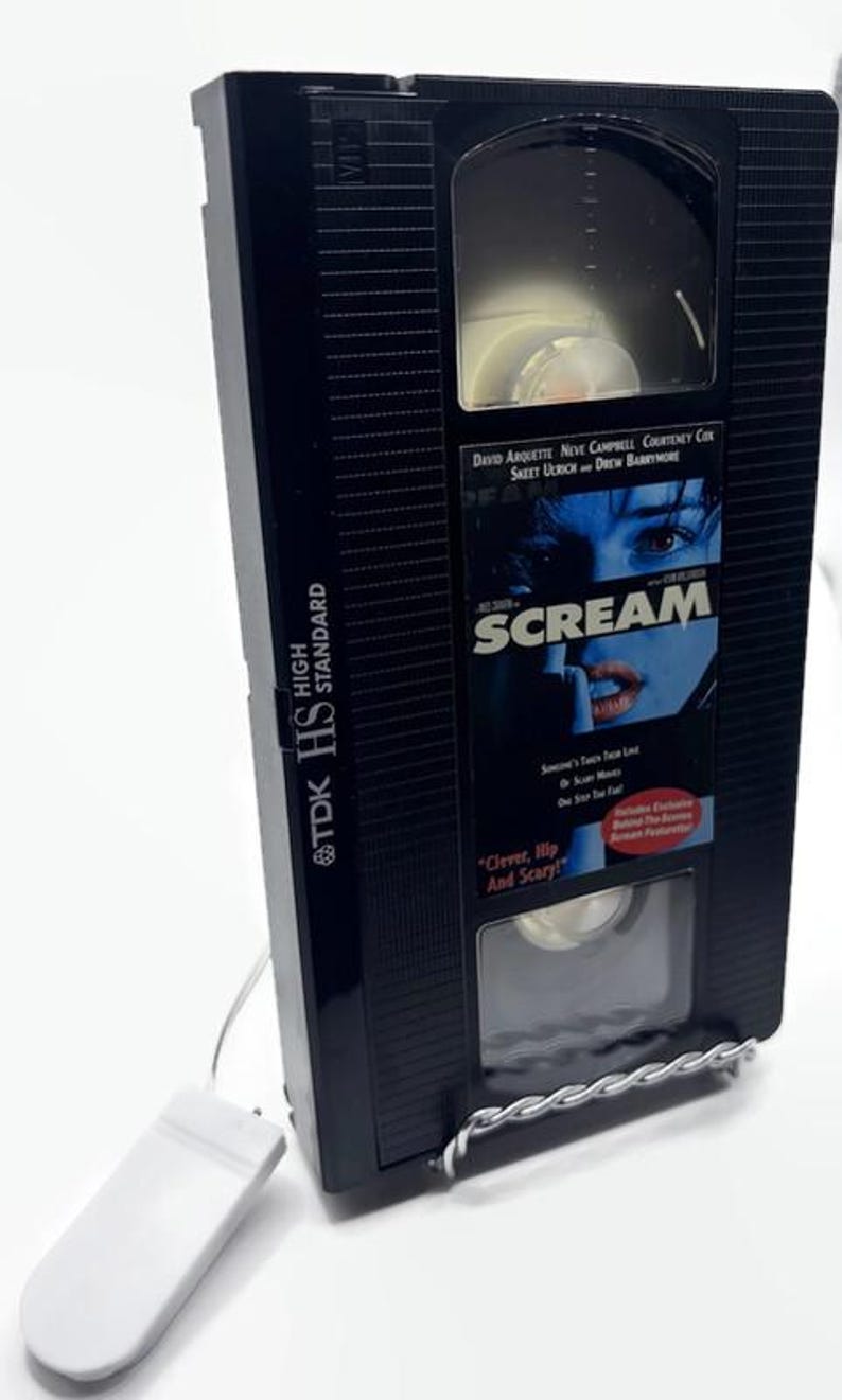 Może przedstawiać: Czarna kaseta VHS z filmem "Scream" z tekstem "Clever, Hip And Scary!" i nazwiskami aktor&oacute;w. Taśma jest trzymana przez srebrny druciany stojak i ma dołączony biały prostokątny przedmiot.