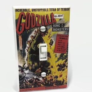 May include: A light switch cover featuring a vintage Godzilla comic book design. The cover has a white toggle switch and two white screws. The artwork includes the text "INCREDIBLE, UNSTOPPABLE TITAN OF TERROR!" and "GODZILLA KING OF THE MONSTERS!"