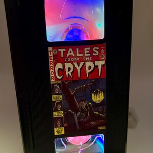 May include: A black VHS tape converted into a lamp, featuring a comic book cover from "Tales From The Crypt." The lamp has a clear plastic front with colorful LED lighting. The comic cover shows a ghoulish figure in a graveyard.