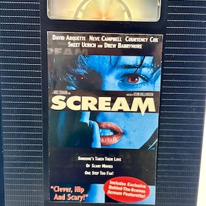 Może przedstawiać: Czarna kaseta VHS z filmem grozy "Scream" z Davidem Arquette, Neve Campbell i Courteney Cox. Okładka przedstawia niebieski obraz twarzy i ust, z tytułem filmu pogrubioną czcionką. Taśma zawiera naklejkę z napisem "Includes Exclusive Behind-The-Scenes Scream Featurette!"