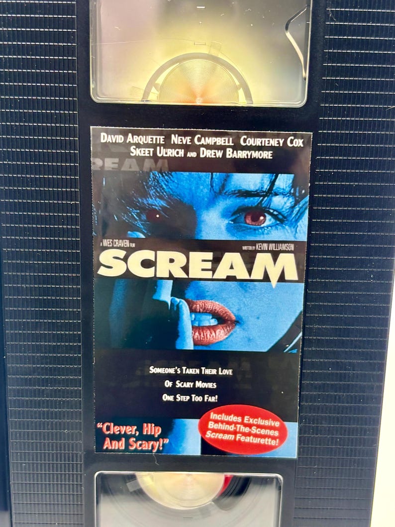 Może przedstawiać: Czarna kaseta VHS z filmem grozy "Scream" z Davidem Arquette, Neve Campbell i Courteney Cox. Okładka przedstawia niebieski obraz twarzy i ust, z tytułem filmu pogrubioną czcionką. Taśma zawiera naklejkę z napisem "Includes Exclusive Behind-The-Scenes Scream Featurette!"