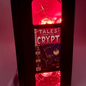 May include: A black, retro-style lamp with red internal lighting. The lamp features a comic book cover from "Tales from the Crypt" and a silver metal base. The top and bottom sections of the lamp have a circular design.